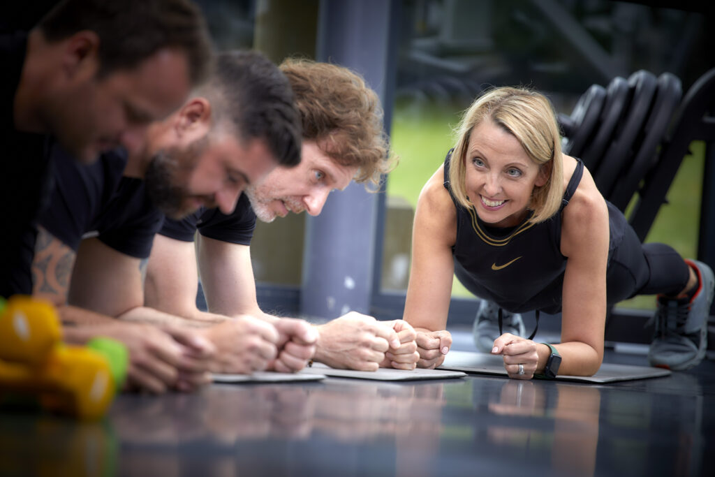 Personal trainer encouraging a small group holding plank exercises during a gym workout.