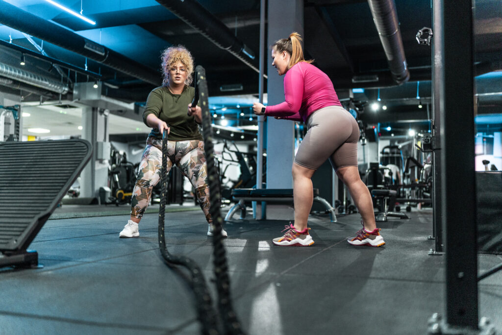 Two women doing a strength workout with battle ropes in a gym during a guided training session.
