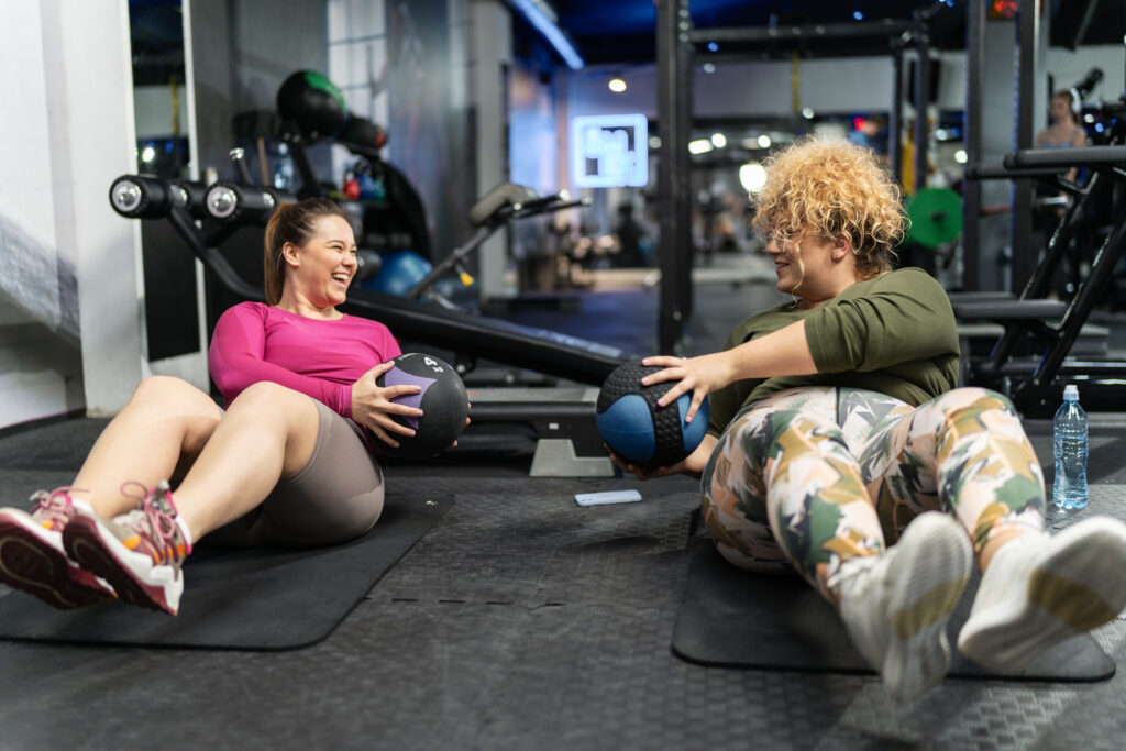 Two women exercising with medicine balls in a gym during a supportive fitness session.