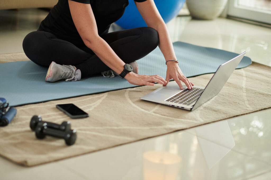 Person sitting on a yoga mat at home using a laptop to follow an online workout, with dumbbells nearby.