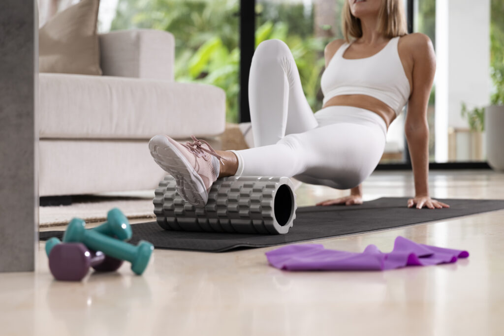 Person foam rolling on a yoga mat at home with dumbbells and a resistance band nearby.