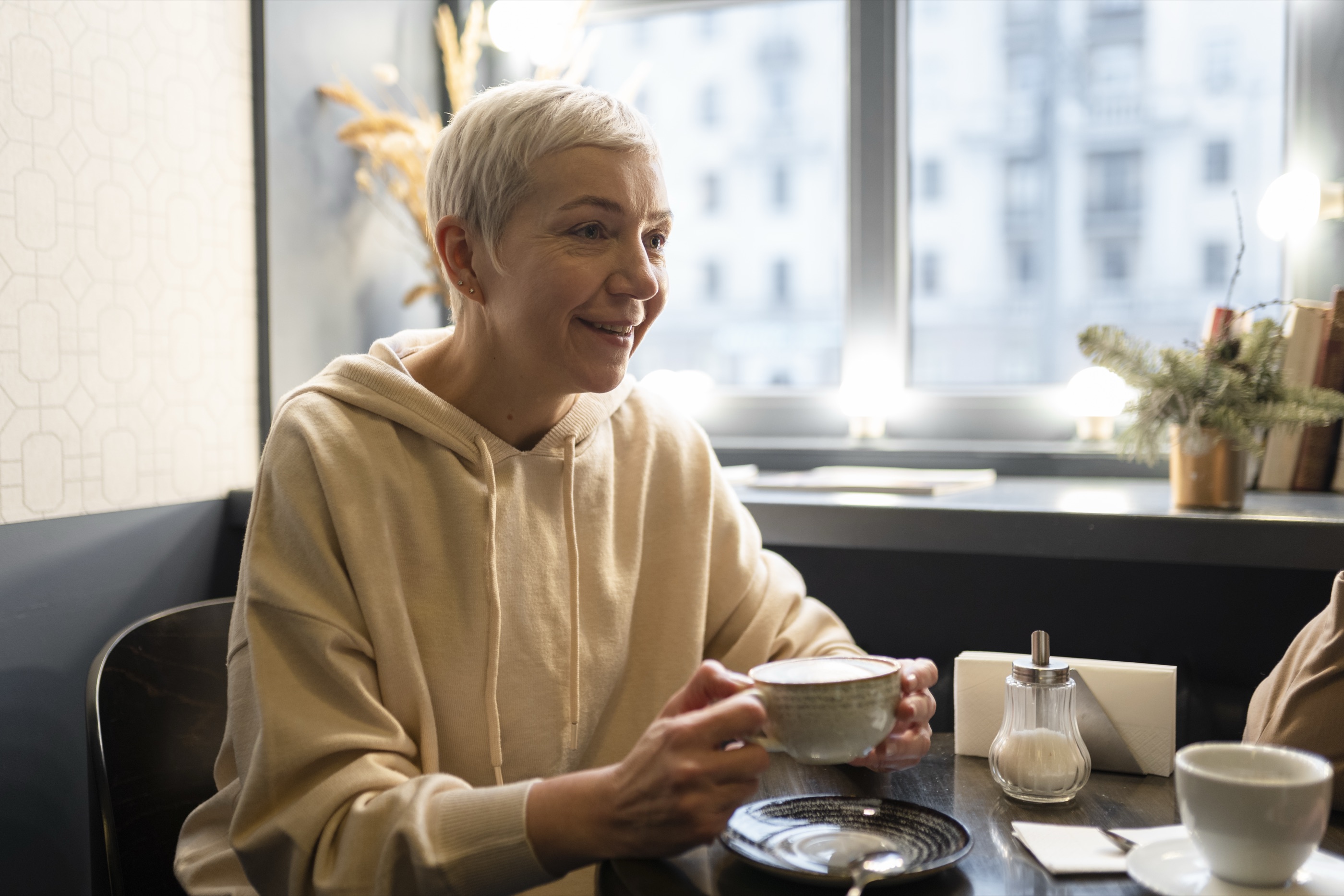 Woman in her fifties with short grey hair smiling over a coffee in a cafe.