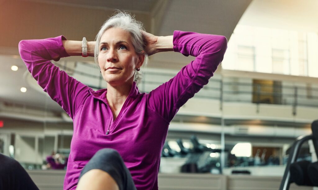 Woman with grey hair doing sit-ups in a gym, hands behind her head.