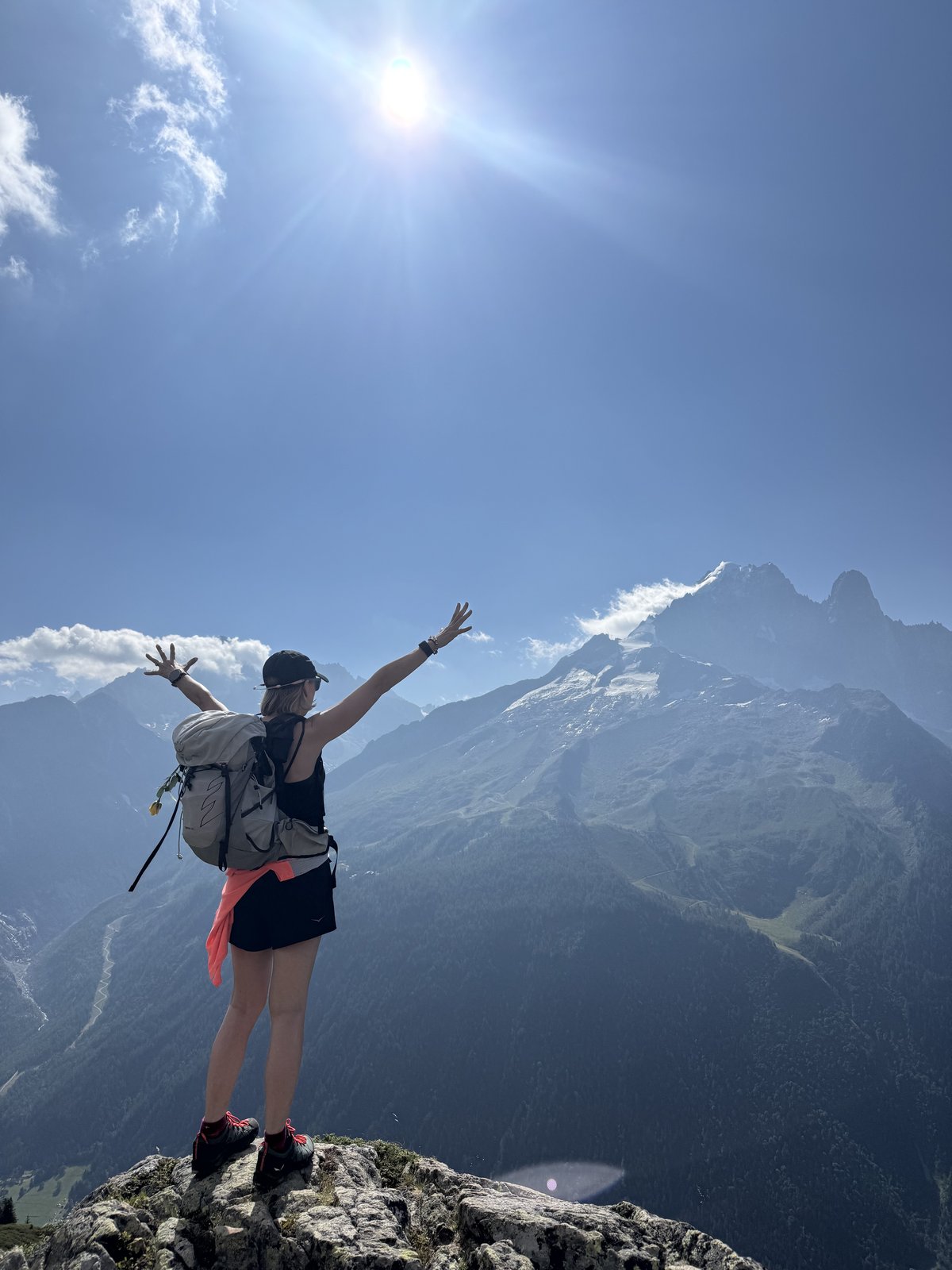 Tracey Pinfold standing on a rocky summit with arms outstretched, the Mont Blanc massif behind her